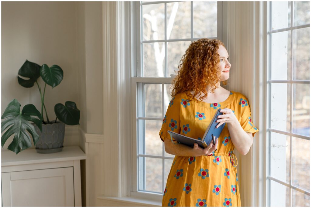 Image from branding photoshoot for Bethany Collins Consulting shows a girl with red curly hair looking out a large window with a blue notebook in hand.